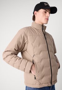 Beige pufferjacka med hög krage, dragkedja framtill och två sidfickor. Har en kviltad textur och diskret branding på kragen.