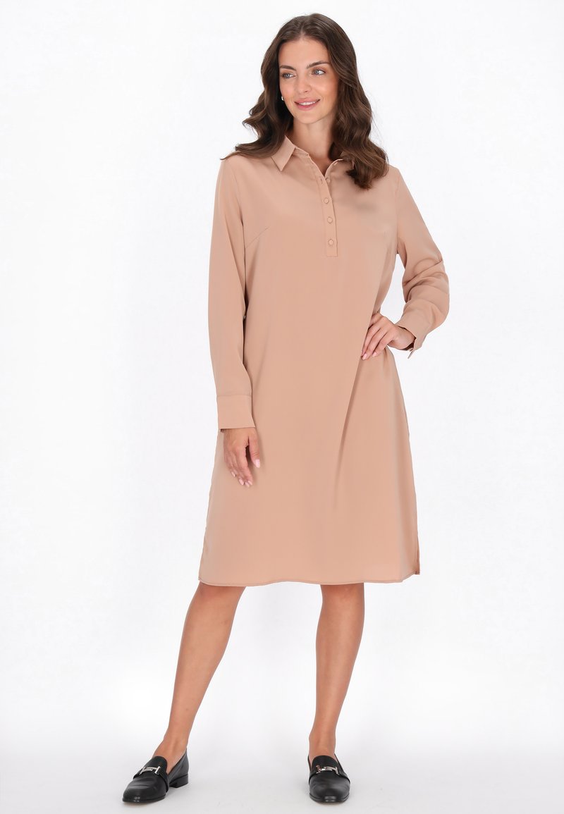 Beiges langärmliges Kleid mit Kragen und durchgehender Knopfleiste. Glattes Material mit geradem Schnitt und seitlichen Schlitzen. Trägt man mit schwarzen Slippern.