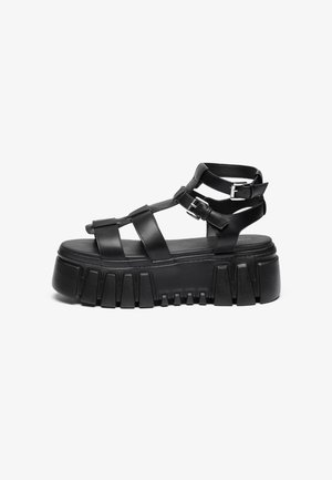 Sort platformsandal med tyk hælsål, tre frontremme og to justerbare ankelremme med spænder.