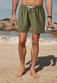 Rohelised ujumispüksid elastse vöökohaga ja valge paelaga. Omavad siledat tekstuuri ja ulatuvad kuni reite keskele, taustal on ranna vaade.