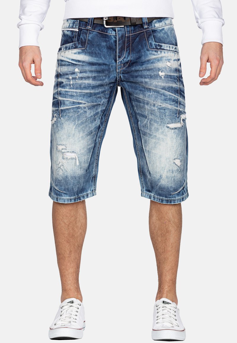 Shorts en denim bleu clair avec des détails déchirés, ourlet effiloché, et cinq poches. Portés avec des baskets blanches sous un haut à manches longues blanc.