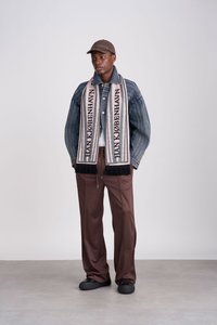 Jeansjakke med vertikale striper, svart og beige skjerf med frynser, brune bukser, svarte sko og en brun caps. Enkelt design og nøytrale farger.