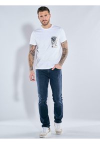 White cotton t-shirt with a front pocket featuring a black and brown abstract pattern, paired with dark blue slim-fit jeans and light sneakers.