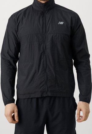 Man draagt een zwarte New Balance lichtgewicht jas met rits en zwarte broek, staand met ontspannen armen langs zijn zij.
