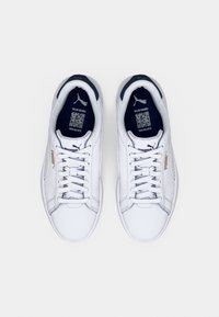 Pár bílých tenisek Puma s tmavě modrým vnitřkem, bílými tkaničkami a zlatým logem Puma na bocích, zobrazený zeshora na bílém pozadí.