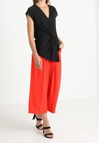 Svart, kortärmad topp i omslagstyp med knutet detalj, i kombination med lösa, ljusröda, vida byxor och svarta sandaler med ankelrem.