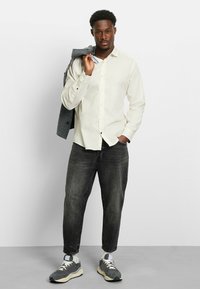 Mann in weißem Hemd mit Knöpfen und schwarzen Jeans hält eine graue Jacke über der Schulter, steht auf einem grauen Boden vor einer weißen Wand.