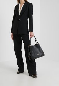 Nainen, joka on pukeutunut mustaan pant suit -pukuun ja valkoiseen paitaan, pitää kädessään mustaa nahkalaukkua, jossa on kultaiset yksityiskohdat, seisten valkoista taustaa vasten.