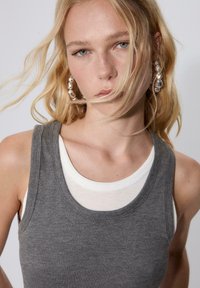 Grauer, gerippter Tanktop, über einem weißen, eng anliegenden Oberteil getragen. Die Accessoires umfassen große, metallic-farbene Creolen. Die Haare sind wellig und zerzaust.