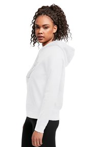 Vit hoodie med en framficka och snörning, med en enkel grafisk design. Modellen bär den tillsammans med svarta leggings, vilket visar passform och tygstruktur.