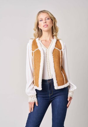 Blondine die een beige shearling-vest draagt over een witte blouse met lange mouwen en kanten manchetten en donkerblauwe spijkerbroek, staand tegen een effen achtergrond.