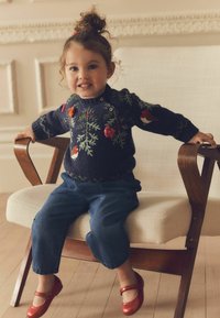 Marineblauer Strickpullover mit Blumen- und Vogelstickerei, kombiniert mit Jeanshosen und roten glitzernden Schuhen, auf einem Holzstuhl sitzend.