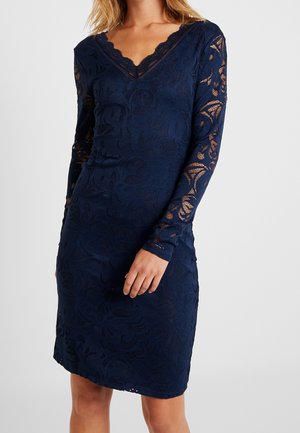 Woman wearing a navy blue, long-sleeve lace dress with a V-neckline and floral patterns, standing against a light gray background.