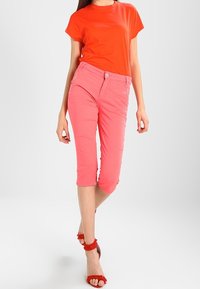 Světlé červené tričko v kombinaci s krátkými růžovými kalhotami. Kalhoty mají zapínání na knoflík a přední kapsy. Celý vzhled dotvářejí červené sandály na řemínky.