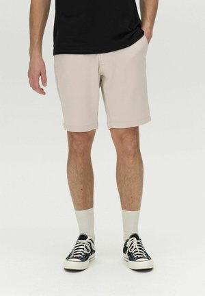 Briși din bumbac bej cu marginile rulate, asociați cu teniși negri și șosete albe până la glezne, purtați cu un tricou negru. Look casual.