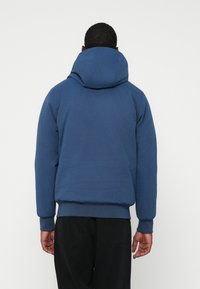 Man in een blauwe gewatteerde hoodie en zwarte broek, staand met zijn rug naar de camera tegen een witte achtergrond.