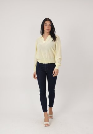 Kobieta z długimi ciemnymi włosami stoi na jednolitym tle, ubrana w jasnżółtą bluzkę z długim rękawem, ciemne obcisłe jeansy oraz białe sandały na obcasie z odkrytymi palcami.