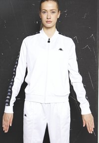 Weiße Trainingsjacke mit Reißverschluss vorne, schwarze Akzente an den Ärmeln, mit einem Logo auf der Brust und gemusterten Details entlang der Arme.