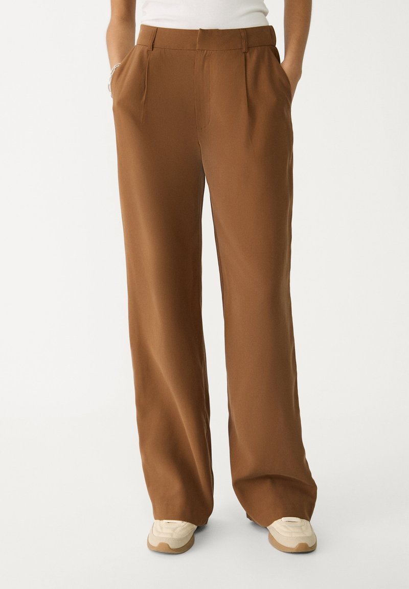 Pantalons larges marron avec une texture lisse, dotés de poches avant et d'une coupe ajustée. Associés à des chaussures de sport de couleur claire.