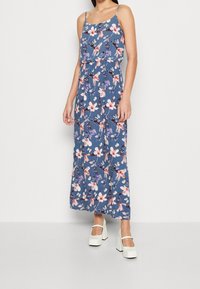 Robe maxi florale avec des fines bretelles, fond bleu, motif de fleurs roses et violettes, et taille élastique. Associée à des chaussures à talons bloc blanches.