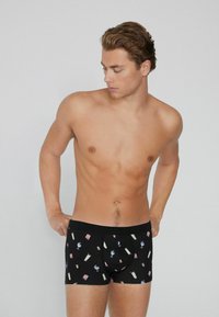 Joven ajustando calzoncillos boxers negros con patrones de cócteles coloridos, de pie frente a un fondo gris claro liso.