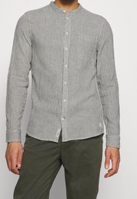 Gestreiftes Hemd mit Knöpfen in Schwarz und Weiß, mit langen Ärmeln, einem Mandarin-Kragen und einer Brusttasche. Der Stoff ist strukturiert und leicht.