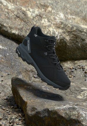 Botte de randonnée Columbia noire avec semelle texturée flottant au-dessus d'un terrain extérieur rocheux.