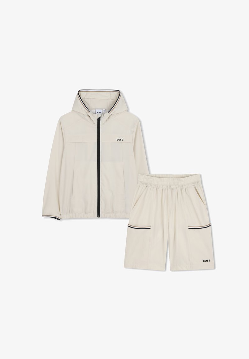 Ensemble veste et short beige clair, en tissu lisse. La veste est dotée d'une capuche, d'une fermeture éclair à l'avant, de deux poches et d'accents noirs. Le short possède des poches latérales.