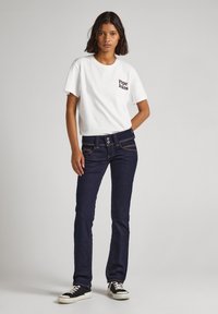 Balta trumpomis rankovėmis marškinėliai su "Pepe Jeans" logotipu, derinami su tamsiai mėlynomis prigludusiomis džinsinėmis kelnėmis, turinčiomis dvigubas sagas ir matomus siuvimo detales.