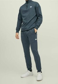 Mörkgrå, texturerad huvtröja med halvdragkedja och logotyppatch, parat med matchande avsmalnande joggers. Vita sneakers fulländar outfiten.