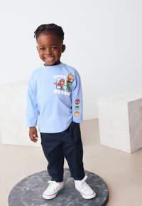 Chemise bleu clair à manches longues avec des graphiques de super-héros, associée à un pantalon foncé et des baskets blanches. Caractérisée par des accents imprimés ludiques sur les manches.