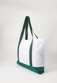 Sac fourre-tout blanc et vert avec un corps en maille texturée, un fond vert uni et des poignées en tissu vert. Présente une forme rectangulaire adaptée au transport.