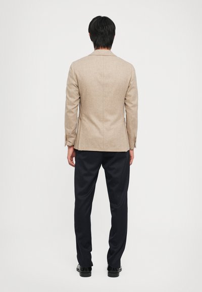 Beige maßgeschneiderter Blazer mit einem Schlitz, strukturiertem Stoff und Knopfakzenten. Kombiniert mit dunkelblauen Hosen und schwarzen Schuhen, von hinten betrachtet.