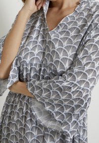 Klänning med flare-ärmar i lätt tyg, med en blå och svart fläktmönster på en ljus bakgrund. V-ringning och rynkad midjedetalj.