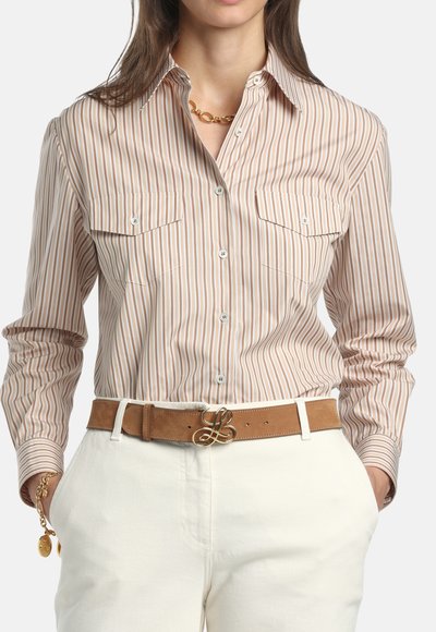 Žena u bež i bijeloj košulji s okomitim prugama na kopčanje, bijelim hlačama, smeđem remenu s zlatnom kopčom i zlatnim nakitom.