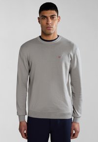 Suéter de manga larga gris claro de material suave, con cuello redondo y un pequeño emblema rojo y azul en el pecho.