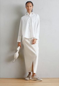 Camisa branca de botões com mangas longas, combinada com uma saia lápis branca com uma fenda frontal e ténis bege; modelo segura uma bolsa.