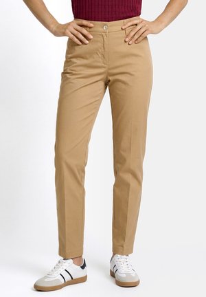 Frau trägt beige eng anliegende Hose, burgunderfarbenes Oberteil und weiße Sneakers mit schwarzen Streifen, steht mit Händen in den Hüften.