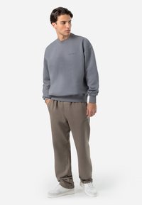 Grå sweatshirt med ribbede manchetter og kant, parret med beige sweatpants. Modellen bærer hvide sneakers og står foran en neutral baggrund.
