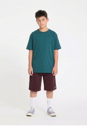 Garçon debout portant un t-shirt bleu sarcelle, un short bordeaux, des chaussettes blanches et des baskets violettes sur un fond blanc.