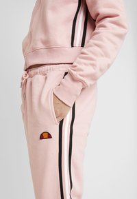 Rosa fleece-träningsoverall med svartvita randiga detaljer, försedda med en sidoficka och en liten broderad logotyp ovanför knät.