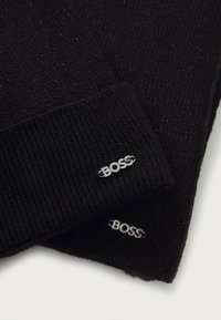 Mustat teksturoitu neulepipo, jossa on kimaltavia lankoja. Pipossa on hopeiset "BOSS"-logot koristeina taittuneessa reunassa ja tiukka muotoilu.