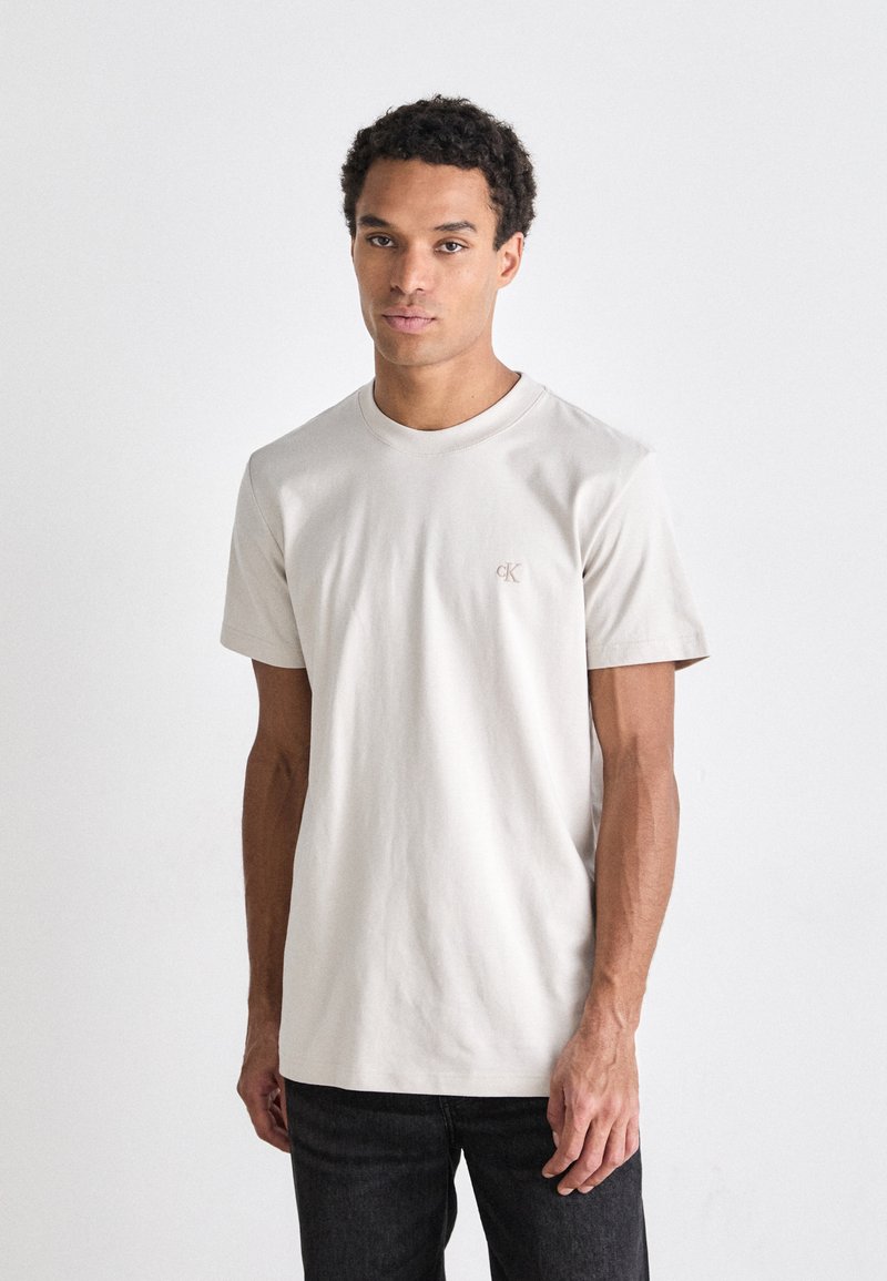 Camiseta color beige claro de algodón suave, cuello redondo, mangas cortas, con un pequeño logo en el pecho. Combinada con jeans oscuros.