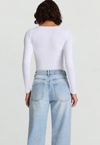 Donna con capelli ricci castani che indossa una maglia bianca aderente a maniche lunghe e jeans a gamba larga color azzurro chiaro a vita alta, rivolta di spalle alla fotocamera.