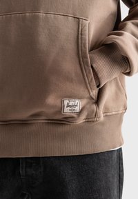 Brown sweatshirt made of cotton blend fabric, featuring a front pocket, ribbed cuffs, and a branded tag on the lower left.