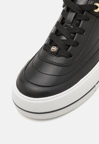 Must musta sneaker, millel on tekstuuriga viimistlus, ümar varvas, mustad paelad ja kuldne logo aktsent. Valge tekstuurne tall lisab kontrasti.