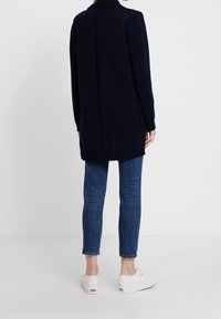 Granatowy długi kardigan z kołnierzem, prążkowanymi mankietami i krawędzią oraz teksturowanym wzorem dzianiny, noszony na niebieskich dżinsach i białych trampkach.