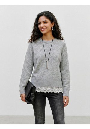 Femme aux cheveux bouclés portant un pull gris et un top en dentelle, tenant une pochette noire, souriante et regardant sur le côté sur un fond uni.