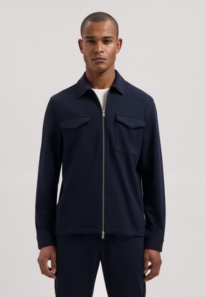 Man draagt een marineblauw jack met rits en twee borstzakken over een wit shirt, staande tegen een effen lichte achtergrond.