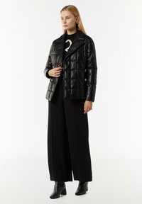 Schwarze, gequiltete Kunstlederjacke mit Reverskragen, doppelten Knöpfen und cropped Schnitt, getragen über einem schwarzen Outfit und Stiefeletten mit Absatz.